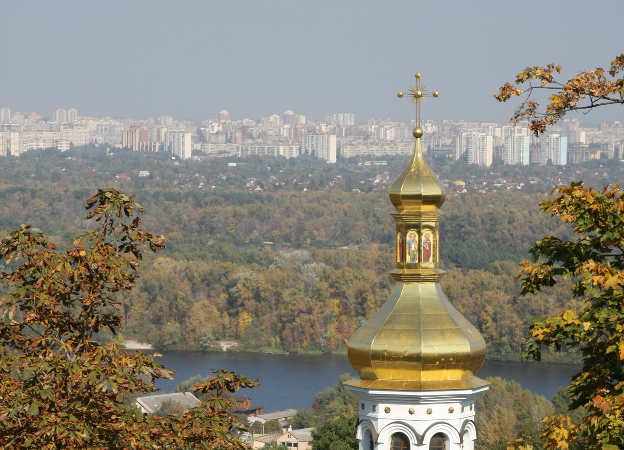 Золотой купол православного храма с осенними листьями на переднем плане и городским пейзажем Киева на заднем фоне