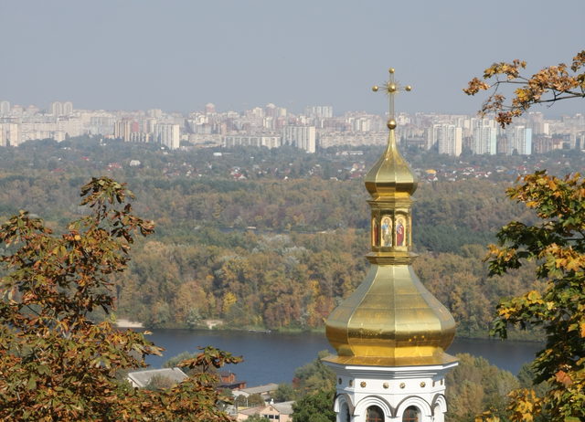Золотой купол православного храма с осенними листьями на переднем плане и городским пейзажем Киева на заднем фоне