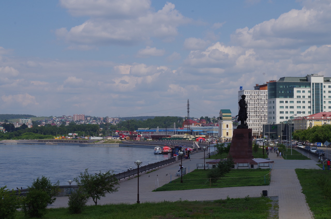 Вид на набережную Ангары в Иркутске с памятником и городскими зданиями на заднем плане.