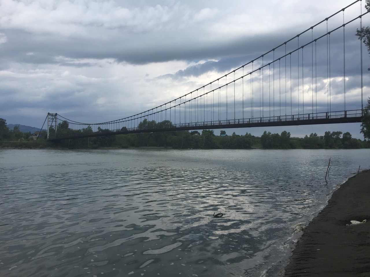 Вид на длинный подвесной мост через широкую реку Катунь с берега.