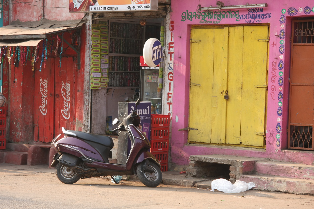 Фиолетовый скутер Hero Activa припаркован на фоне красных и розовых стен магазинов.
